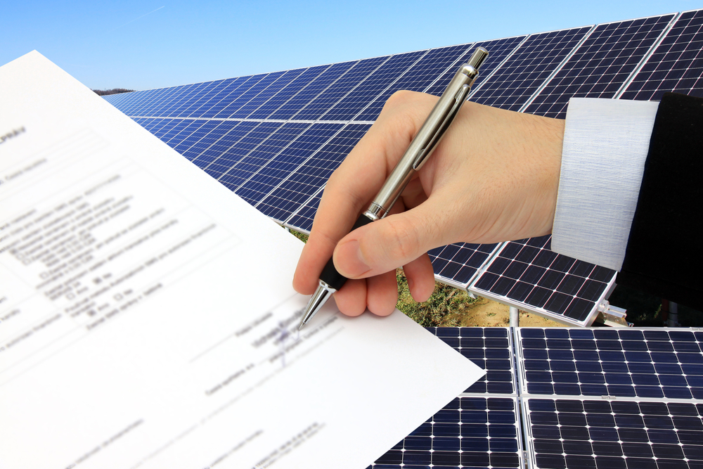 Business professionals signing a contract in front of a solar power station set in green natural surroundings, representing a sustainable energy agreement.