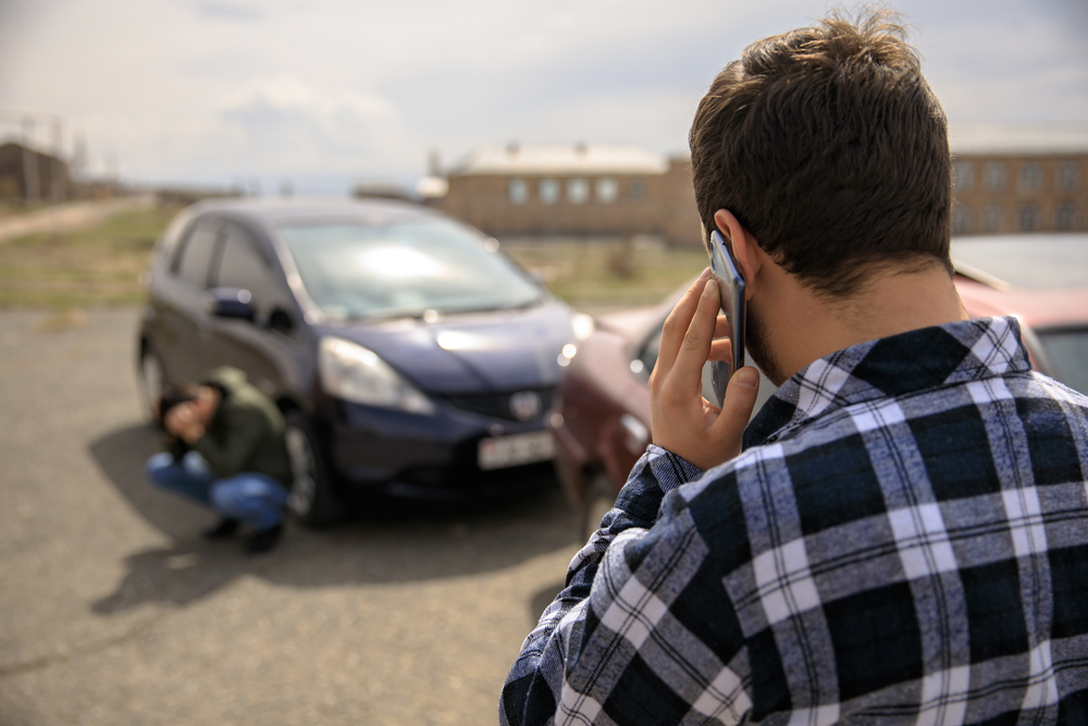 Sad man calling roadside assistance after a car crash, standing near damaged vehicle.