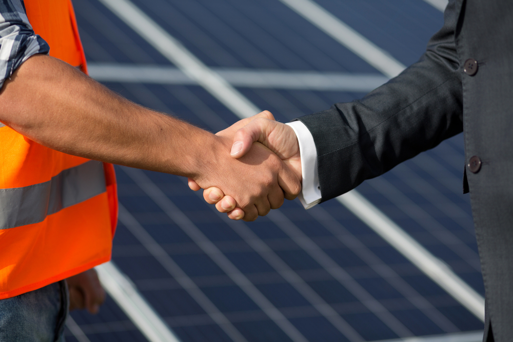 Close-up of two Asian male engineers wearing safety vests shaking hands in front of solar panels, symbolizing a business agreement in renewable and green energy